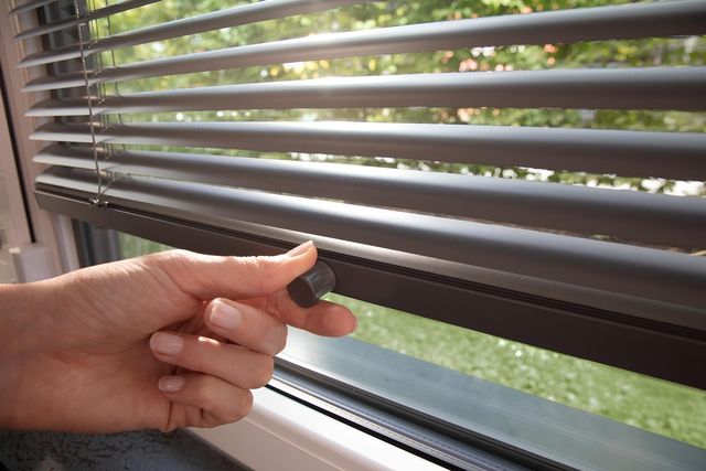 Hand reguliert den Lichteinfall durch Verstellen eines Lamellenbehangs am Fenster