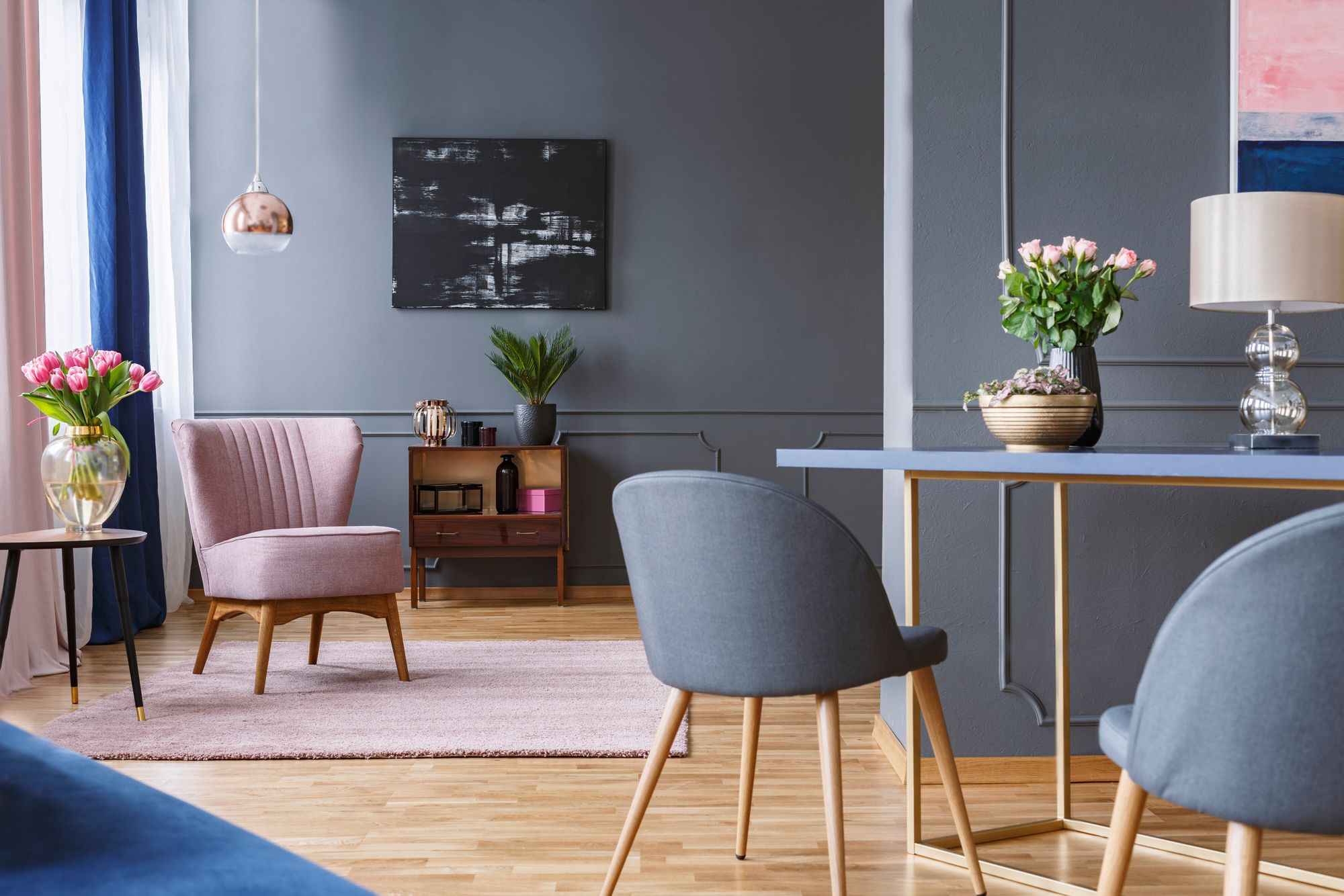Elegant eingerichtetes Wohnzimmer mit rosa Sessel, grauen Wänden, dunklem Sideboard, runder Vase mit Tulpen, Esstisch mit blauer Platte und hellen Holzbeinen.